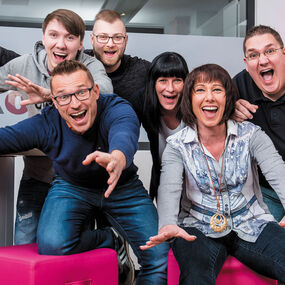 Sechs fröhliche Mitarbeiter eines Telekommunikationsshops posieren lachend im Laden.