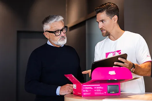 Mann erklärt älterem Kunden Smartphone-Tarif am Tablet, Produktbox auf Tisch